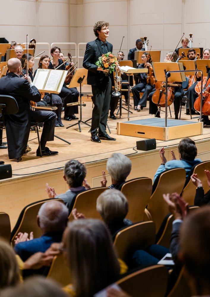 Eloy Pérez Llavata steht mit einem Blumenstrauß in der Hand dem Publikum dankend zugewandt auf der Bühne, das Orchester im Hintergrund