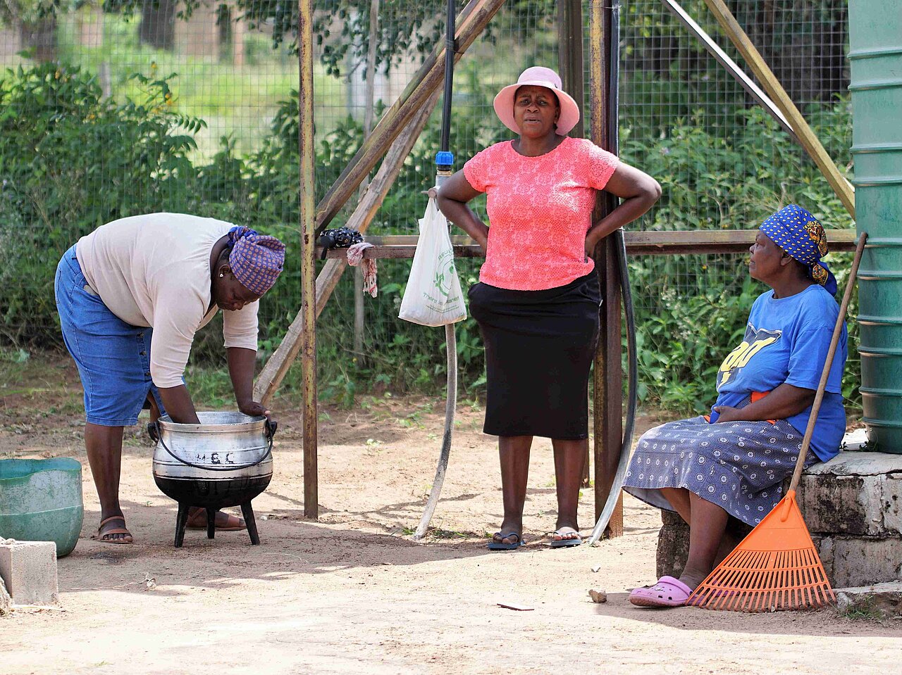 Drei Frauen der Gemeinde Welverdiend in Südafrika sitzen im Schatten, eine Frau reinigt einen Kessel