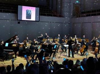 Ein Orchester musiziert auf der Bühne, das Publikum hält Handys mit leuchtendem Display in die Höhe, im Hintergrund ist ein Bildschirm mit Handysymbol