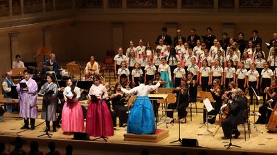 Auf einer Bühne steht ein Kinderchor, Musiker eines Orchesters und eine Dirigentin, im Vordergrund singen vier Sänger in koreanischer Tracht